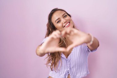 Young hispanic woman standing over pink background smiling in love doing heart symbol shape with hands. romantic concept. 