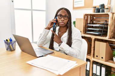 Ofiste çalışan örgülü siyahi bir kadın telefonda konuşuyor. Elleriyle işaret ederek reklamını yapıyor. Ciddi ve sakin bir yüz ifadesi var. 