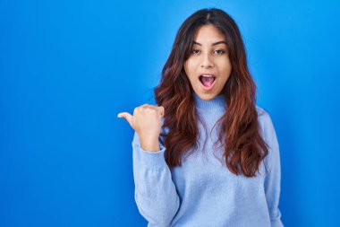 Hispanic young woman standing over blue background surprised pointing with hand finger to the side, open mouth amazed expression. 
