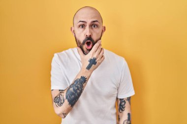 Sarı arka planda dikilen İspanyol dövmeli genç adam elleri çenesinde olan inançsızlıktan, şaşkınlıktan ve şaşkınlıktan büyülenmiş görünüyor. 