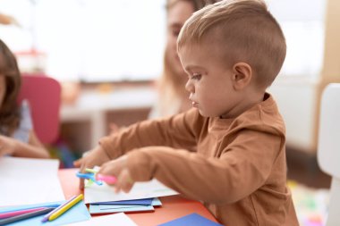 Anaokulunun masasında oturan, not defterine resim çizen sevimli çocuk.