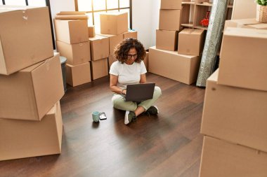 Orta yaşlı İspanyol kadın yeni evinde dizüstü bilgisayar kullanarak gülümsüyor.