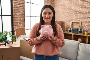 Young hispanic woman smiling confident holding piggy bank at new home