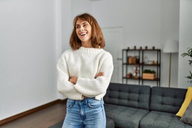 Beyaz bir kadın gülümsüyor. Kollarını kavuşturup evde duruyor.