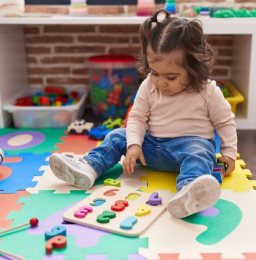 Adorable hispanic girl playing with maths puzzle game sitting on floor at kindergarten