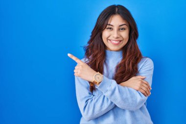 Hispanic young woman standing over blue background with a big smile on face, pointing with hand finger to the side looking at the camera. 