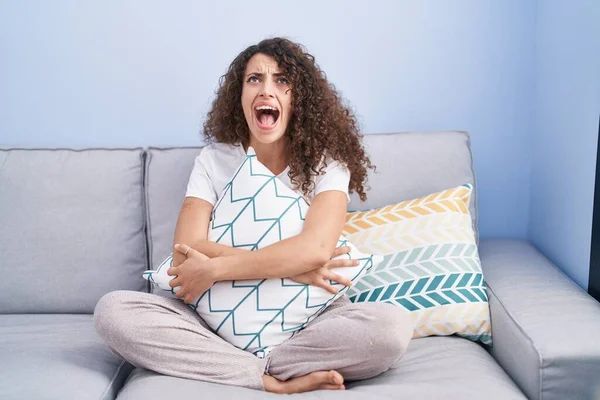 Hispanic woman with curly hair sitting on the sofa at home angry and mad screaming frustrated and furious, shouting with anger looking up. 
