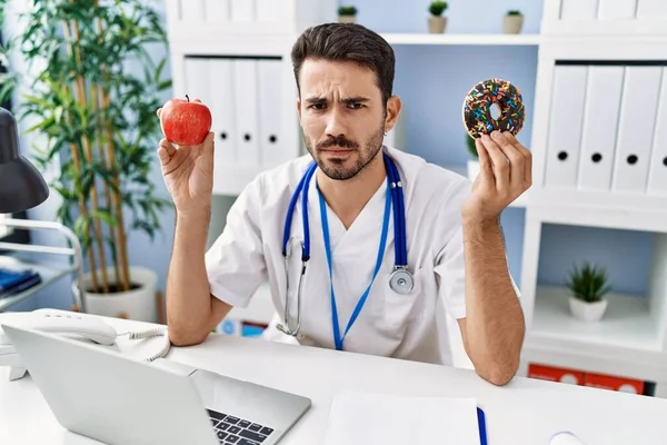 Ung Latinamerikansk Dietist Håller Munk Och Äpple Skeptisk Och Nervös — Stockfoto