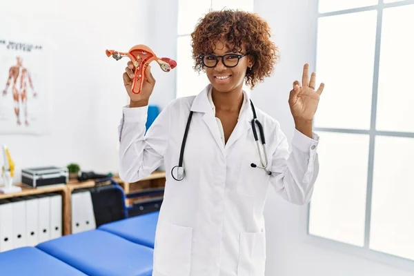 Young african american woman holding anatomical model of female genital organ doing ok sign with fingers, smiling friendly gesturing excellent symbol