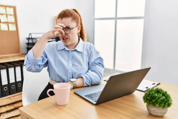 Young redhead woman working at the office using computer laptop smelling something stinky and disgusting, intolerable smell, holding breath with fingers on nose. bad smell
