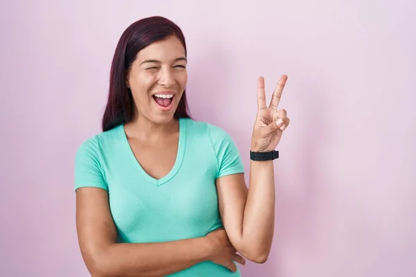 ピンク色の背景の上に立つ若いヒスパニック系の女性は カメラでの勝利サインを指で行うにウィンク幸せな顔で笑っています — ストック写真