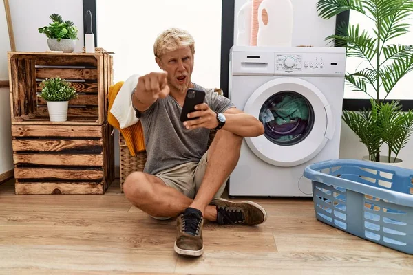 Young Blond Man Doing Laundry Using Smartphone Pointing Displeased Frustrated — Stok fotoğraf