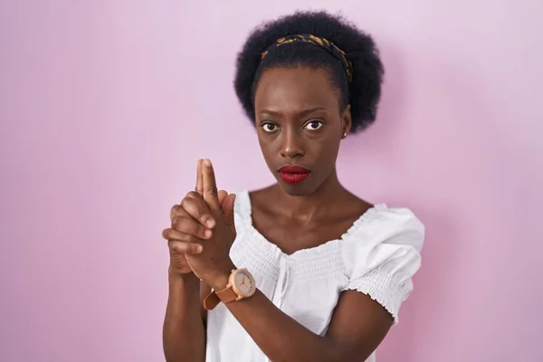 African woman with curly hair standing over pink background holding symbolic gun with hand gesture, playing killing shooting weapons, angry face