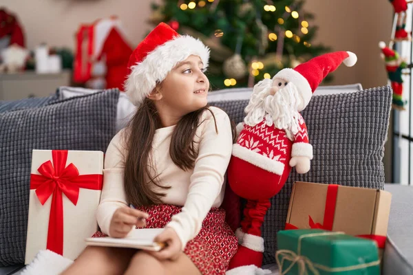 愛らしいHispanic女の子書き込み上のノートブックでソファに座ってクリスマスツリーによって家 — ストック写真