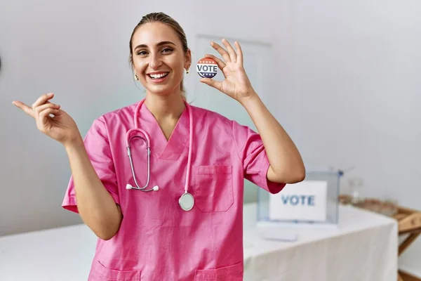 Young nurse woman at political campaign holding usa vote badge smiling happy pointing with hand and finger to the side