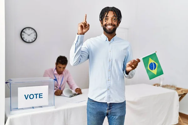 Young hispanic men at political campaign election holding brazil flag surprised with an idea or question pointing finger with happy face, number one