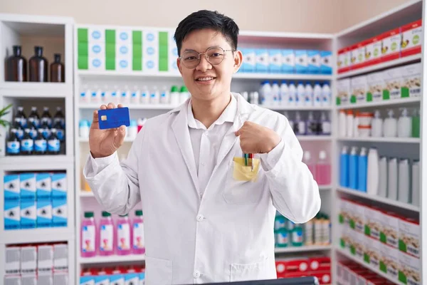Young asian man working at pharmacy drugstore holding credit card pointing finger to one self smiling happy and proud