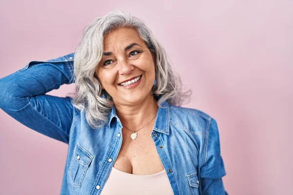 Middle age woman with grey hair standing over pink background smiling confident touching hair with hand up gesture, posing attractive and fashionable
