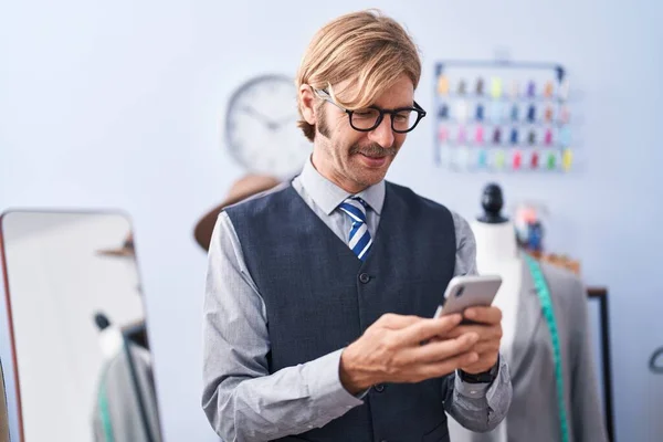 Young blond man tailor smiling confident using smartphone at clothing factory