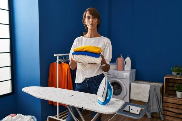 Brunette woman holding folded laundry after ironing making fish face with mouth and squinting eyes, crazy and comical.