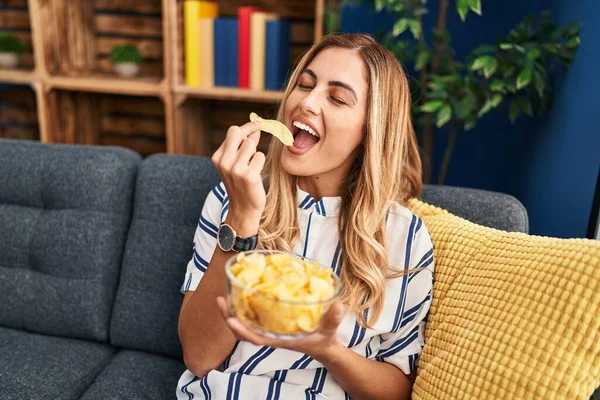 Young Blonde Woman Eating Chips Potatoes Sitting Sofa Home — Stockfoto