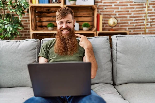 一个留着长胡子的红头发男人坐在客厅的沙发上 用笔记本电脑指着一个快乐而自豪的人 — 图库照片