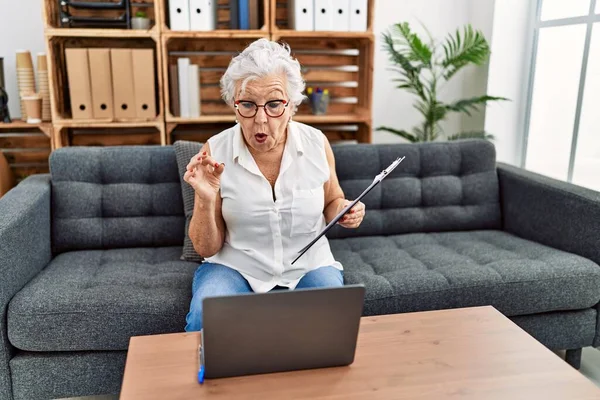 Muayenehanede çalışan gri saçlı yaşlı bir kadın çevrimiçi terapi yapıyor korkmuş ve ağzı açık bir şekilde şaşırmış, inançsız bir yüz ifadesi var. 
