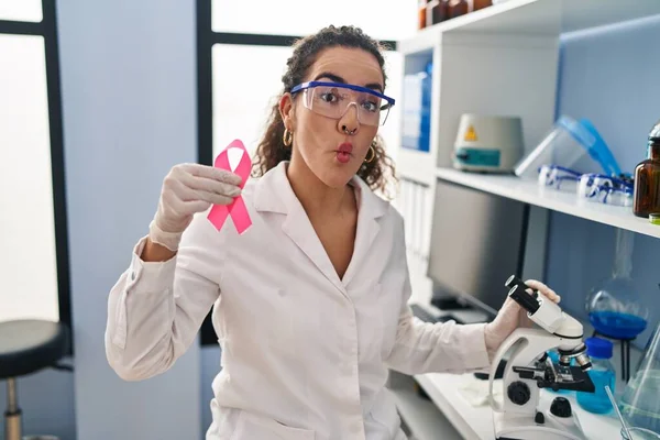 Young hispanic woman working at scientist laboratory looking for breast cancer cure making fish face with mouth and squinting eyes, crazy and comical.