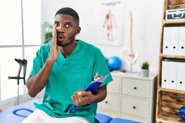 Young african american man working at pain recovery clinic hand on mouth telling secret rumor, whispering malicious talk conversation