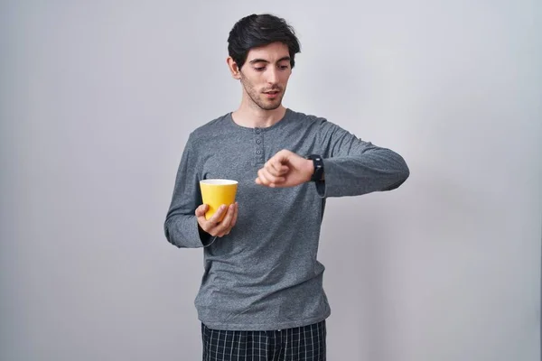 Young hispanic man wearing pajama drinking a cup of coffee looking at the watch time worried, afraid of getting late