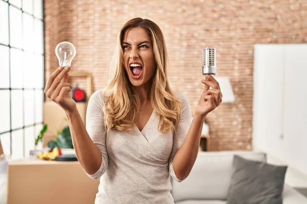 Young blonde woman holding led lightbulb and incandescent bulb angry and mad screaming frustrated and furious, shouting with anger looking up.