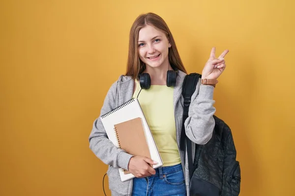 Young caucasian woman wearing student backpack and holding books smiling with happy face winking at the camera doing victory sign with fingers. number two.