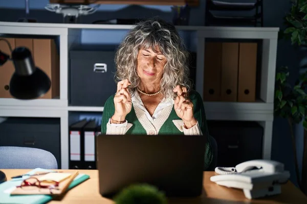 Middle age woman working at night using computer laptop gesturing finger crossed smiling with hope and eyes closed. luck and superstitious concept.