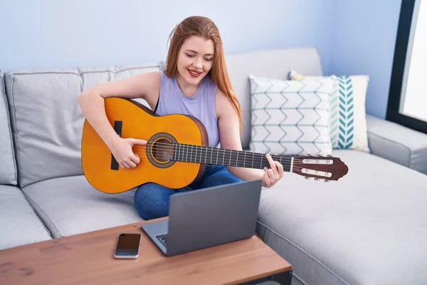 Young redhead woman having online classical guitar class sitting on sofa at home