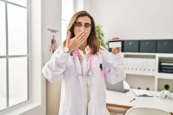 Young doctor woman holding credit card at the clinic covering mouth with hand, shocked and afraid for mistake. surprised expression