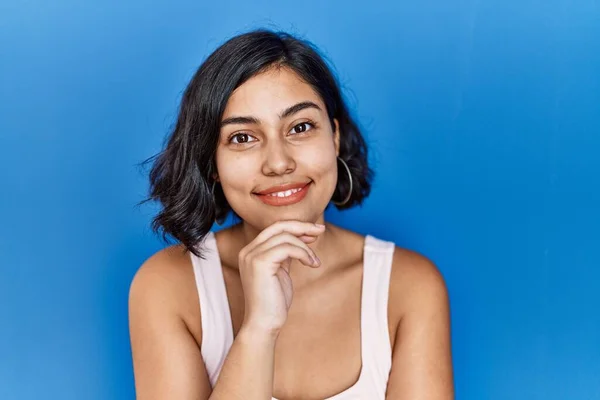 Young hispanic woman standing over blue background looking confident at the camera smiling with crossed arms and hand raised on chin. thinking positive.