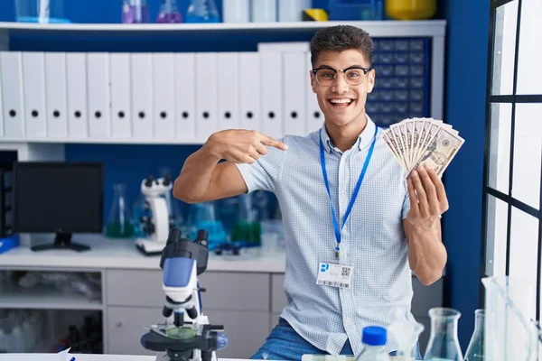 Young Hispanic Man Working Scientist Laboratory Holding Dollars Pointing Finger — Stock Photo, Image