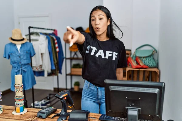 Young hispanic woman working as staff at retail boutique pointing with finger surprised ahead, open mouth amazed expression, something on the front