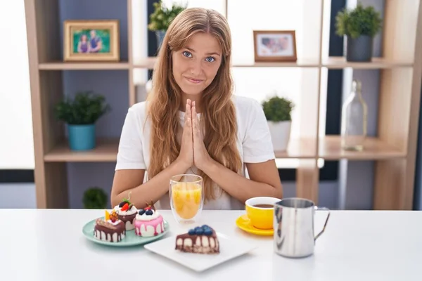 Young Caucasian Woman Eating Pastries Breakfast Praying Hands Together Asking — Stock Fotó