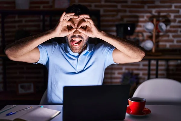 Hispanic man with beard using laptop at night doing ok gesture like binoculars sticking tongue out, eyes looking through fingers. crazy expression.