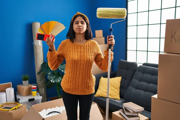 Young hispanic woman painting home walls with paint roller puffing cheeks with funny face. mouth inflated with air, catching air.