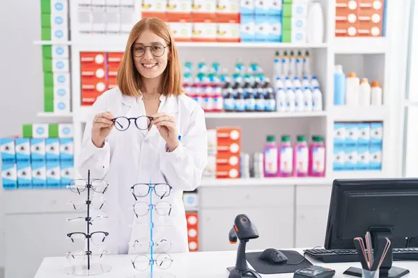 Young Redhead Woman Working Pharmacy Drugstore Holding Glasses Smiling Happy — 图库照片