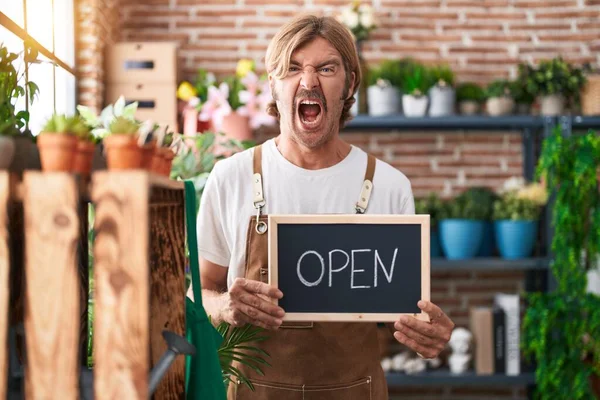Caucasian man with mustache working at florist holding open sign angry and mad screaming frustrated and furious, shouting with anger. rage and aggressive concept.