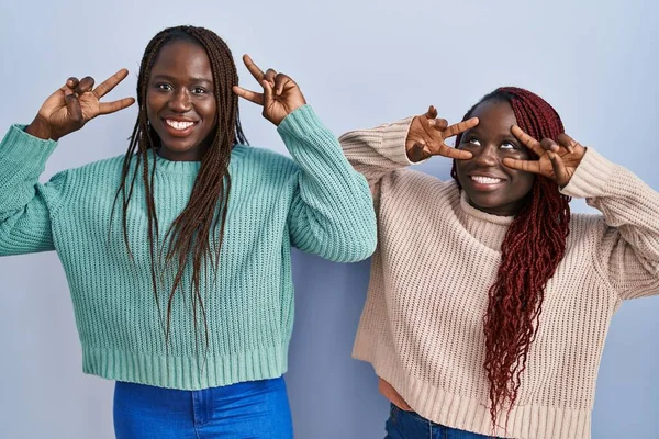 顔の上に指で平和のシンボルをやって青い背景に立っている2人のアフリカの女性 勝利を示す明るい笑顔 — ストック写真