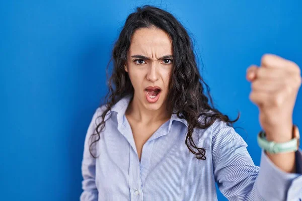 Young brunette woman standing over blue background angry and mad raising fist frustrated and furious while shouting with anger. rage and aggressive concept.