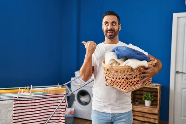 Young Hispanic Man Beard Holding Laundry Basket Pointing Thumb Side — Zdjęcie stockowe