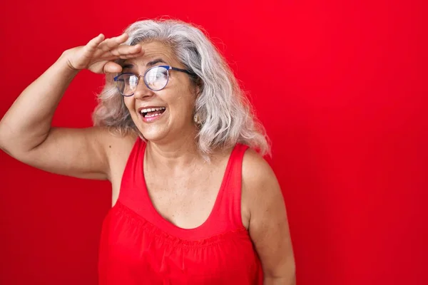 赤い背景の上に立っている灰色の髪を持つ中年の女性は非常に幸せと遠くに手を頭の上に見て笑顔 検索の概念 — ストック写真