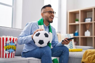 Young hispanic man football hooligan holding ball supporting team smiling and laughing hard out loud because funny crazy joke. 