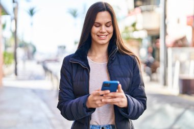 Genç, güzel, İspanyol bir kadın sokakta akıllı telefon kullanarak gülümsüyor.
