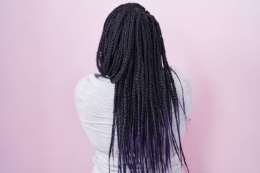 Genç Afrikalı bir kadın pembe arka planda dikiliyor. Arkaya bakıyor ve kollarını kavuşturuyor. 